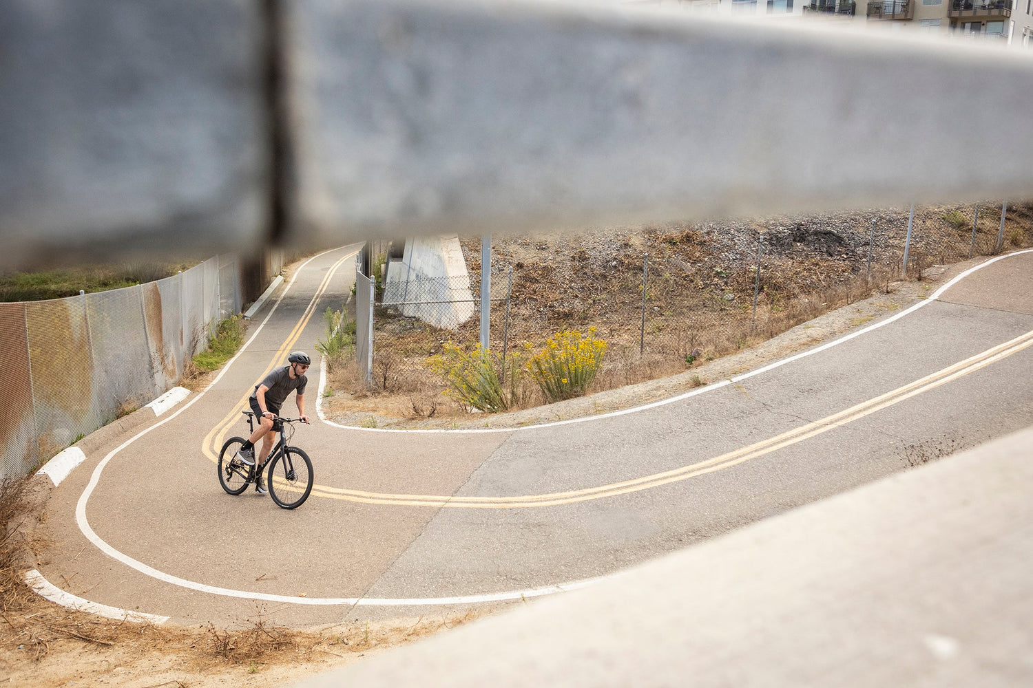A cyclist rides on a winding road surrounded by dry vegetation and metal barriers. The perspective is through a gap in a concrete or metal structure, framing the scene with an urban and somewhat industrial backdrop.