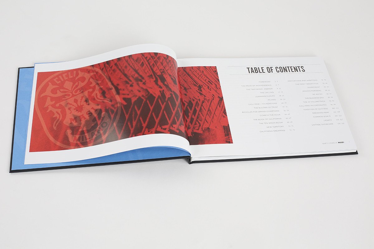 The book 90 YEARS OF MASI by Masi Bikes features a left page with a red abstract design and a right page titled Table of Contents with chapters like Bicycle History and Cicli Masi, resting on a white surface and recounting tales of European bicycle racing.