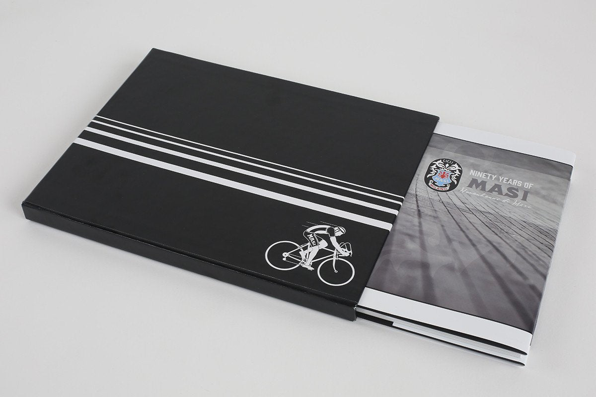 A black rectangular folder with white stripes and a cyclist graphic reveals a booklet titled 90 YEARS OF MASI by Masi Bikes, encapsulating European bicycle racing history, against a plain white background.