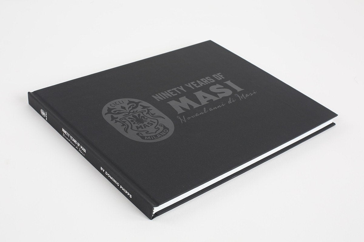 A black hardcover book titled 90 YEARS OF MASI by Masi Bikes rests on a white surface, chronicling bicycle history. Its cover features an emblem and bilingual text (English and Italian), honoring Cicli Masis influence on European bicycle racing.