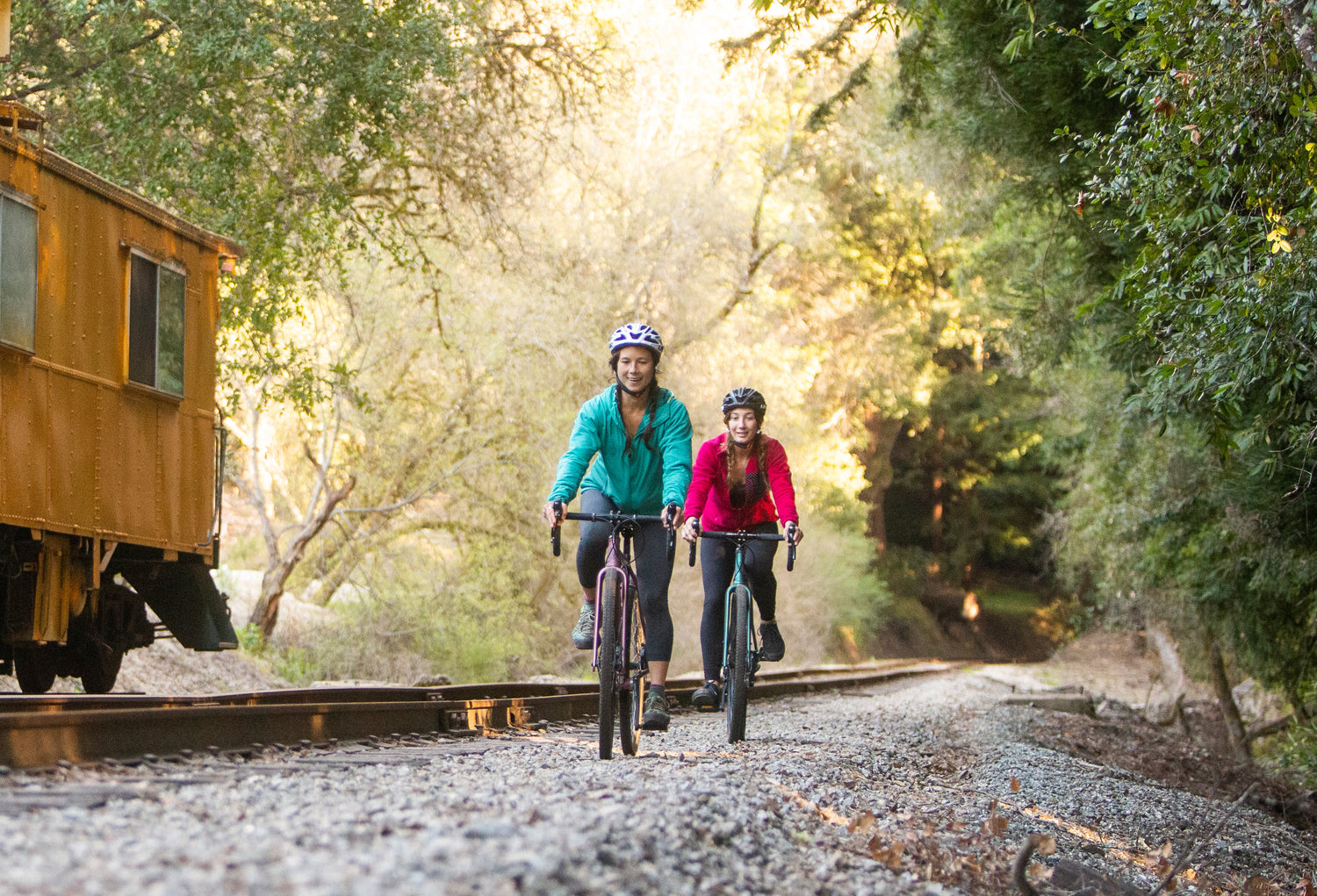 Two cyclists ride along a gravel path beside train tracks in a forested area. They wear helmets and jackets, with sunlight filtering through the trees. A yellow train car is visible on the left.
