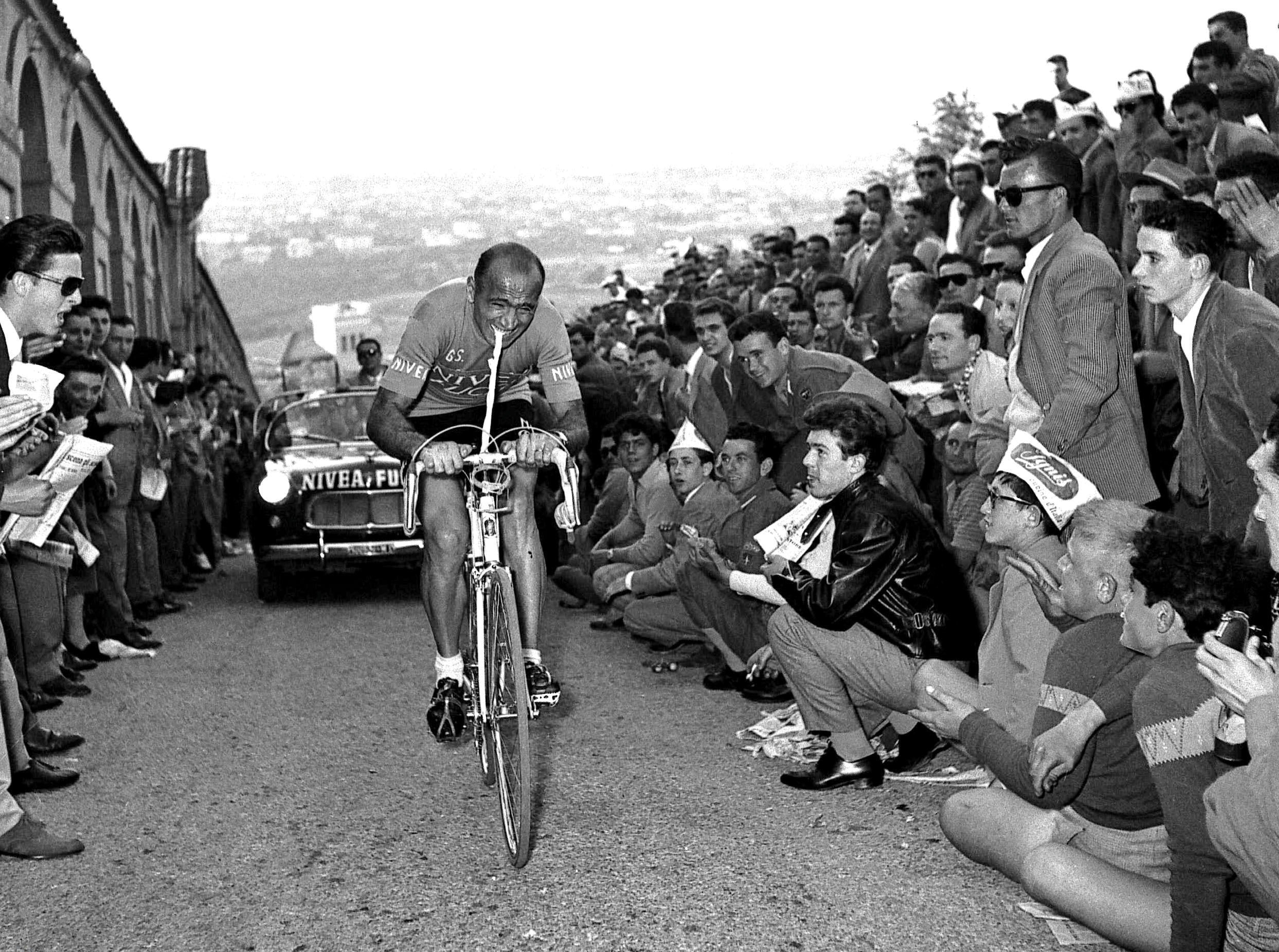 A cyclist in a race rides uphill, surrounded by a crowd of spectators on both sides. The crowd includes men holding newspapers and wearing hats. A support car follows the cyclist closely. The scene conveys excitement and focus.