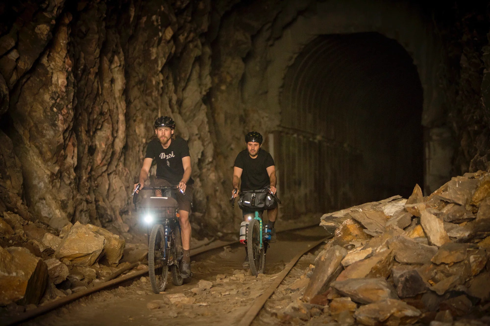 Two cyclists ride through a dimly lit, rocky tunnel. The cyclists, wearing helmets and equipped with lights, navigate a rugged path surrounded by large stones. The tunnels entrance is visible in the background.