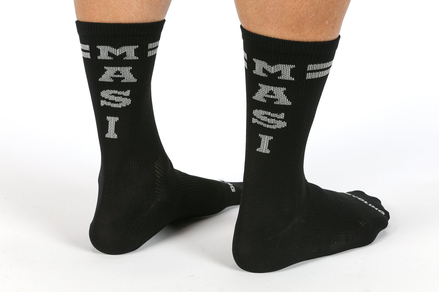 A person wears MASI DNA CYCLING SOCKS BLACK by Masi Bikes, featuring white MASI text on the back. These breathable, sweat-wicking performance socks cover the lower calves, with feet on a white background showcasing their comfort.