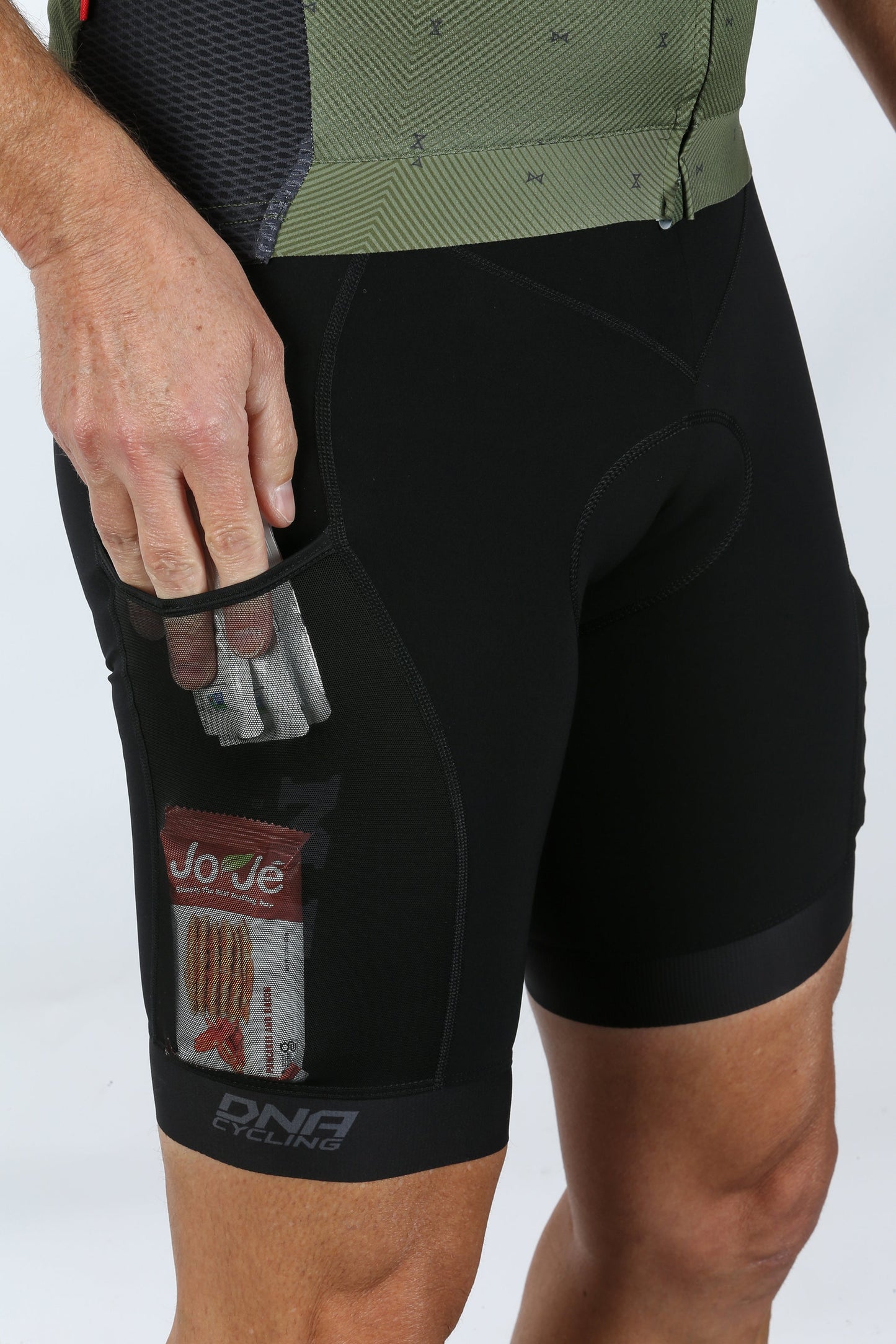 A cyclist in Masi DNA Gravel Bibs, known for ergonomic fit, sports a small transparent pocket with an energy bar. The logo reads DNA Cycling. With their hand beside the pocket, they enjoy optimal comfort on the ride.
