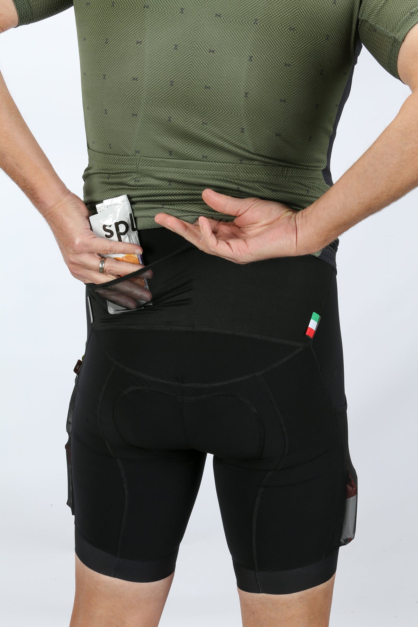 A person wearing a green cycling jersey and MASI DNA GRAVEL BIBS by Masi Bikes is shown from the back. Theyre placing a snack labeled SPL into their ergonomic fit shorts, designed for optimal comfort. The shorts also feature a small Italian flag emblem.