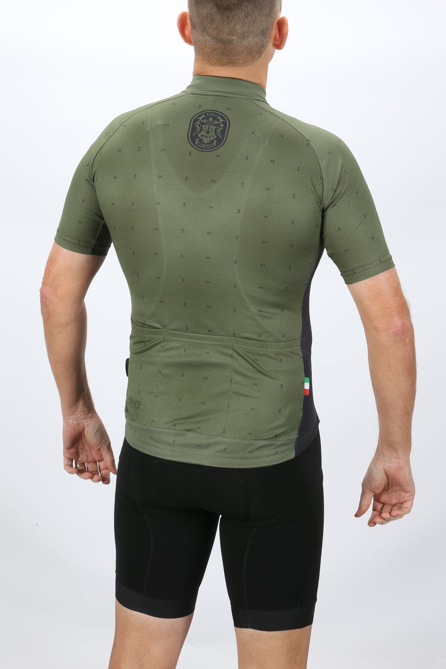 A person in a green MASI DNA RACE JERSEY by Masi Bikes, featuring ultra-aerodynamic short sleeves with a subtle pattern and an Italian flag detail, stands with their back to the camera paired with black cycling shorts.