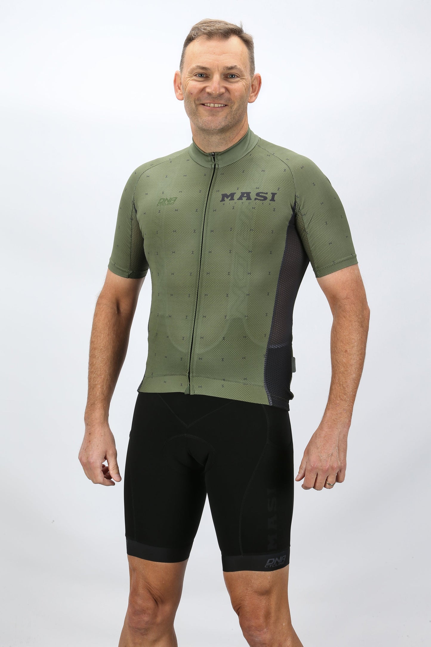 A man wearing a Masi Bikes DNA Race Jersey in green and black cycling shorts, featuring a BIO FIT design, stands smiling against a plain white background.