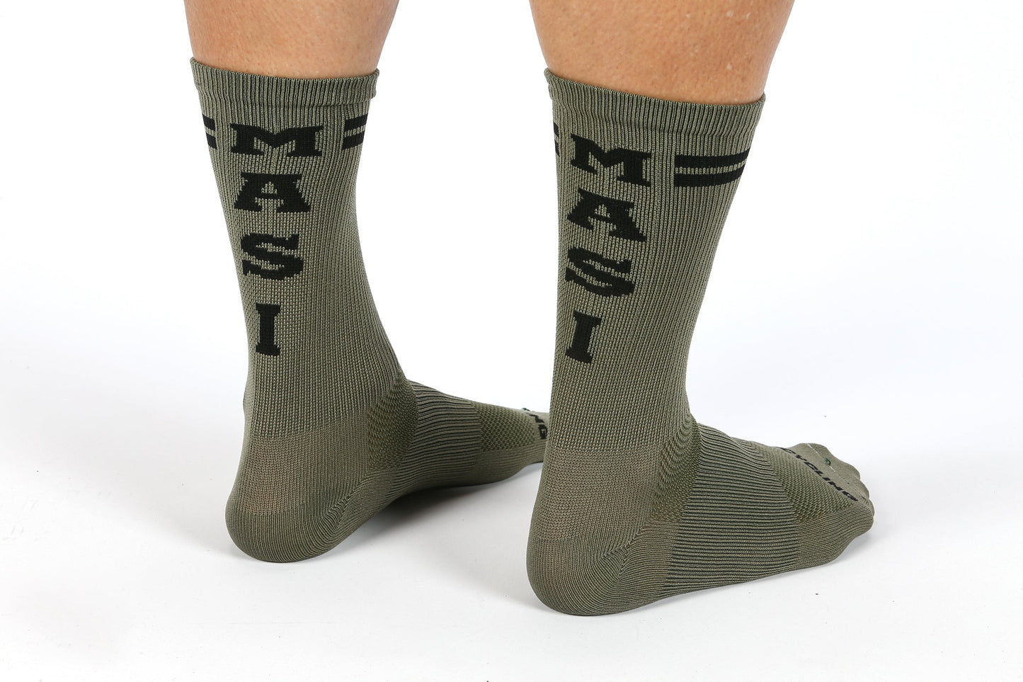 Someone wore MASI DNA cycling socks by Masi Bikes, featuring olive green with MASI in black on the back. The ribbed toes and heels are enhanced by a thin black stripe near the top. These ankle-to-calf coverage socks are breathable and sweat-wicking for all-day comfort.