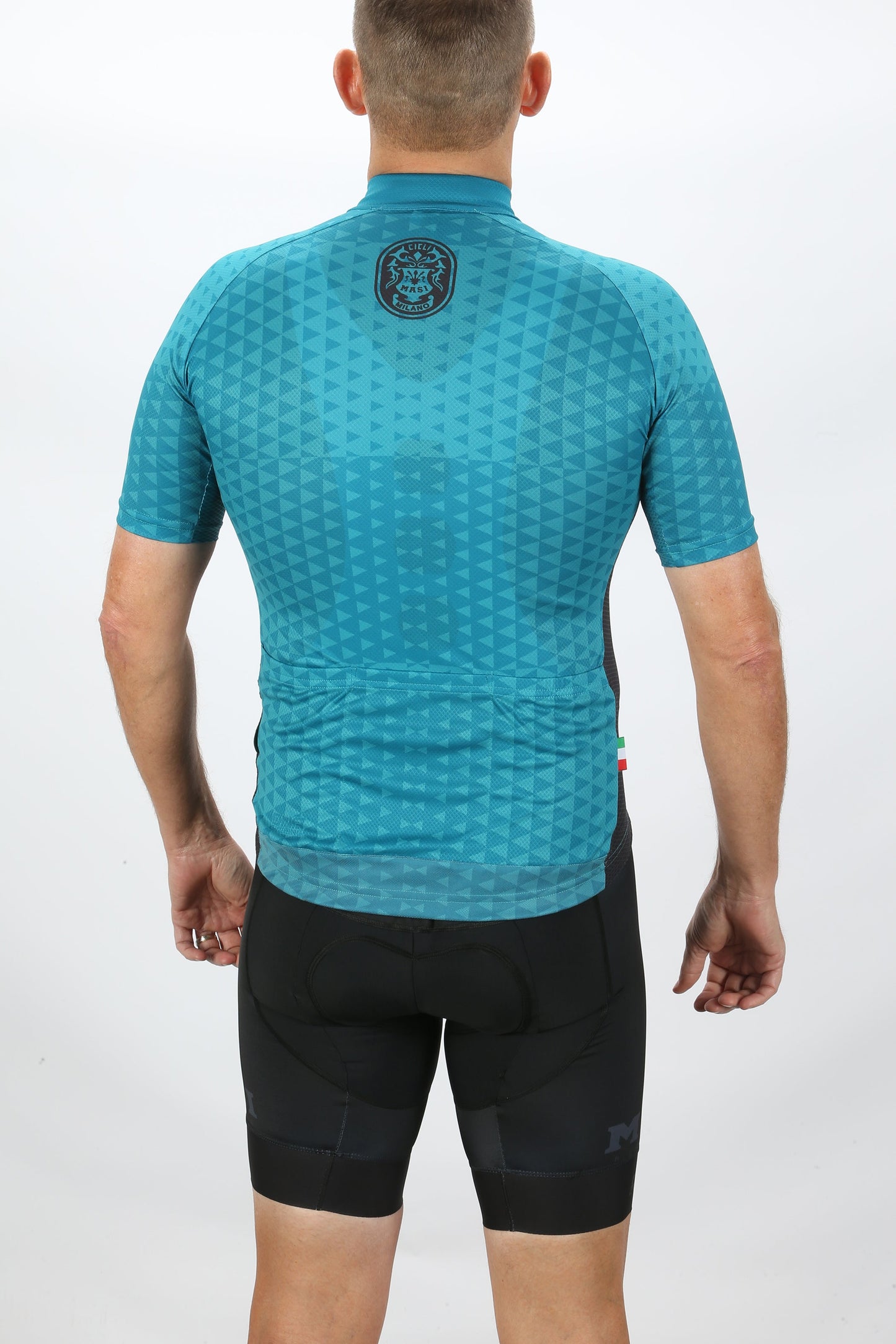 A person in a MASI DNA RACE JERSEY BLUE by Masi Bikes and black shorts faces away. The teal cycling jersey has a geometric pattern, short sleeves, and a small logo near the collar. Engineered with BIO FIT technology, it blends performance with style against the plain white backdrop.