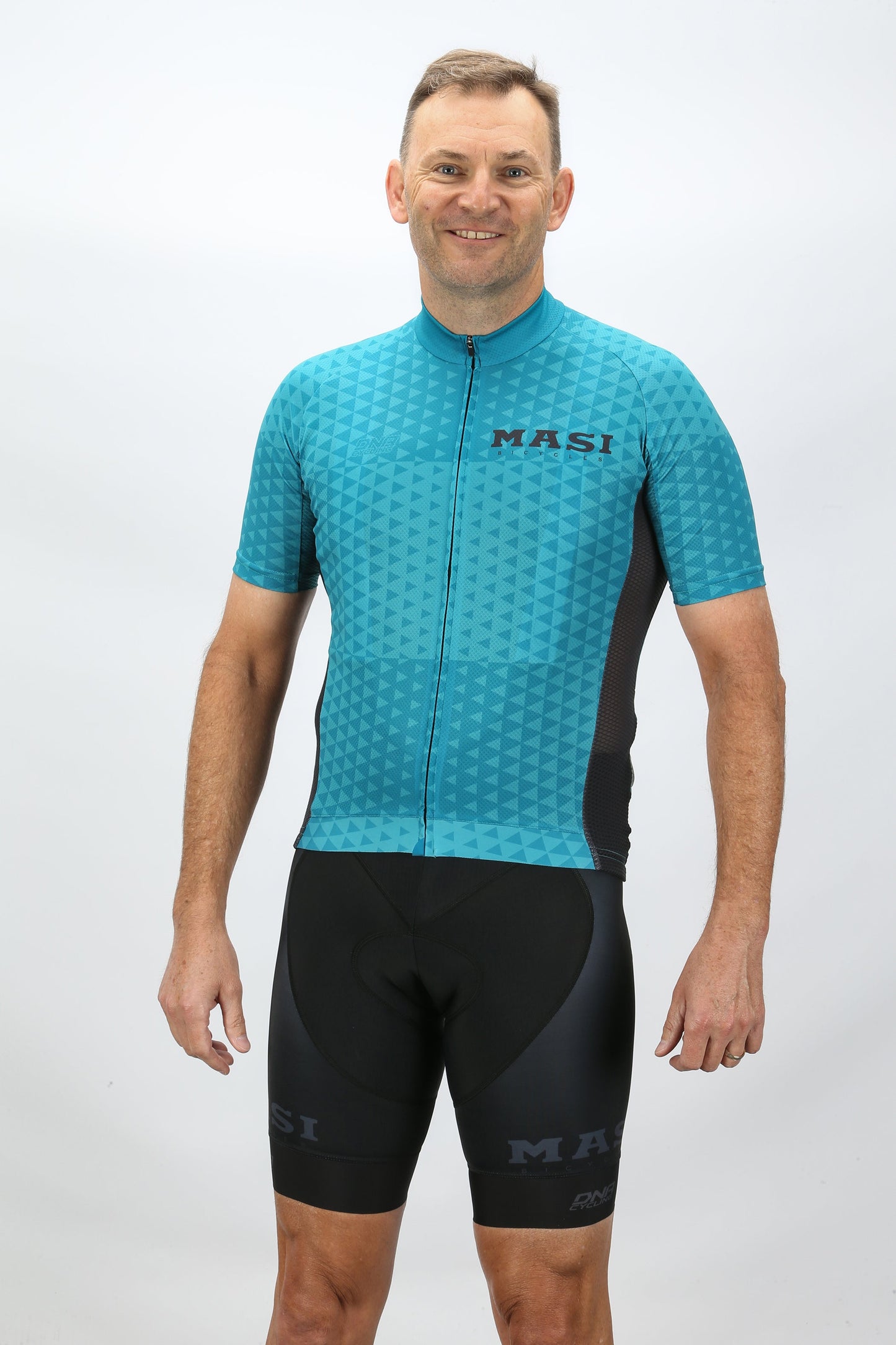 A man wearing a Masi Bikes MASI DNA Race Jersey Blue and black shorts stands smiling against a plain white background.
