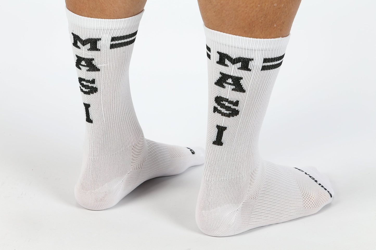 A person stands on a white surface wearing MASI DNA CYCLING SOCKS WHITE by Masi Bikes, featuring the word MASI in bold black letters and two horizontal black stripes near the top. These are sweat-wicking performance socks.