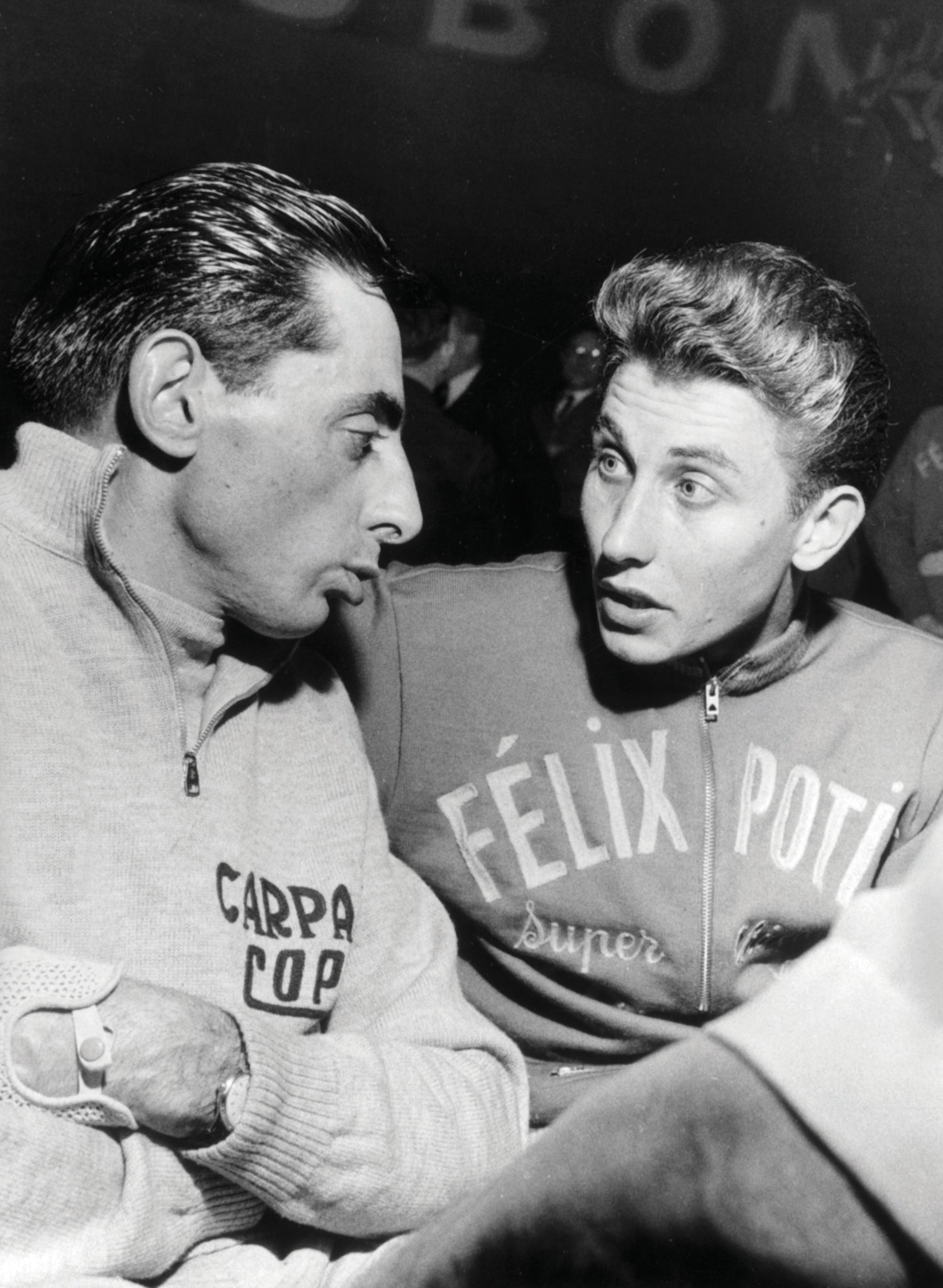 Two cyclists are engaged in conversation, both wearing sports attire with logos. One has a dark slicked-back hairstyle, and the other has light, wavy hair. The setting suggestively involves a sports-related event or discussion.