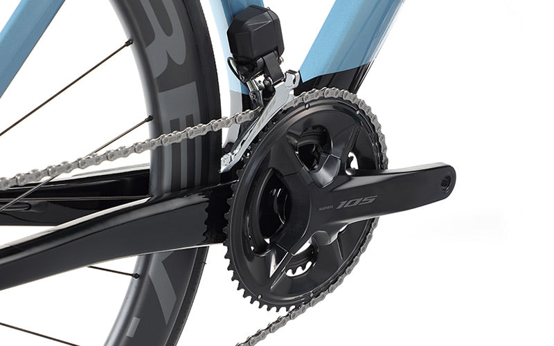 Close-up of a Masi Bikes EVOLUZIONE 1 Carbon road bikes drivetrain, featuring the aerodynamic black Shimano 105 crankset with two chainrings connected to a silver chain. The blue frame complements the rear wheels black spokes and offers a glimpse of the rim.