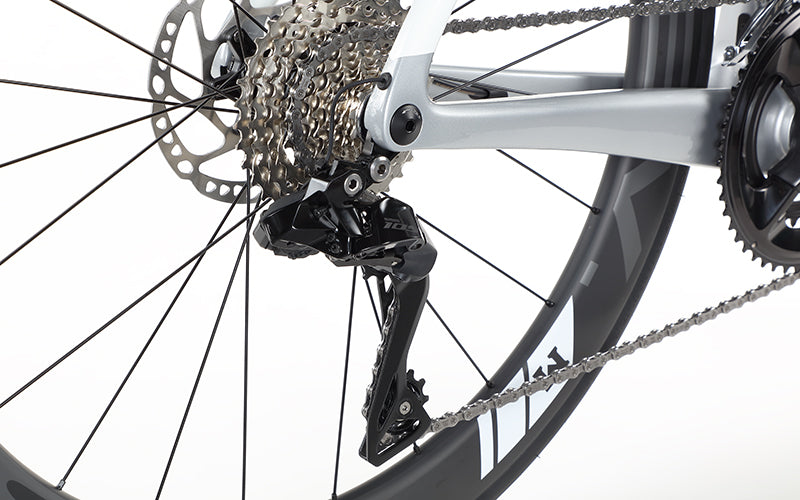 Close-up of the rear derailleur and cassette on a Masi Bikes EVOLUZIONE 1, highlighting its metal gears, chain, and rear wheel spokes against a plain background to emphasize the aerodynamic design and mechanical components.