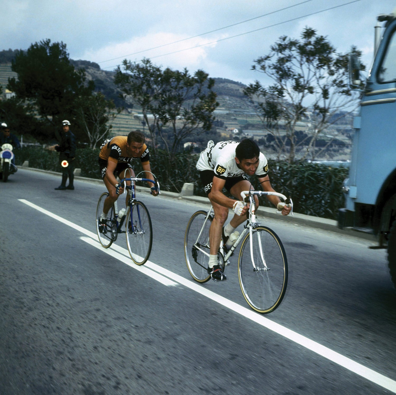 Two cyclists race on a road lined with trees, pursued by a motorcycle. The lead cyclist, in a black and white jersey, is closely followed by another in a yellow and black jersey. An officer and a blue vehicle are visible on the roadside.