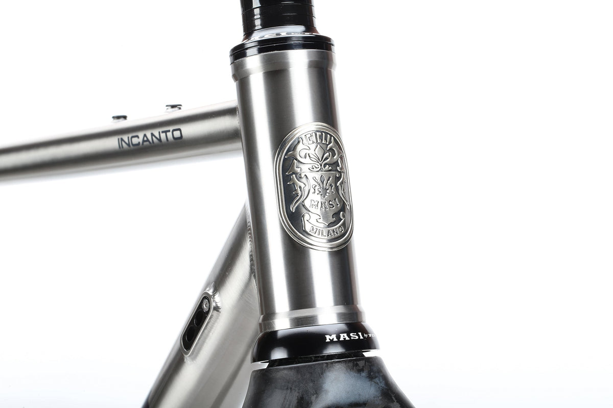 A close-up of the Masi Bikes INCANTO Ti FRAMESET reveals a sleek titanium gravel bike frame. It features an engraved emblem with intricate detailing at the front and INCANTO branded on the polished, metallic 3AL/2.5V tubing.