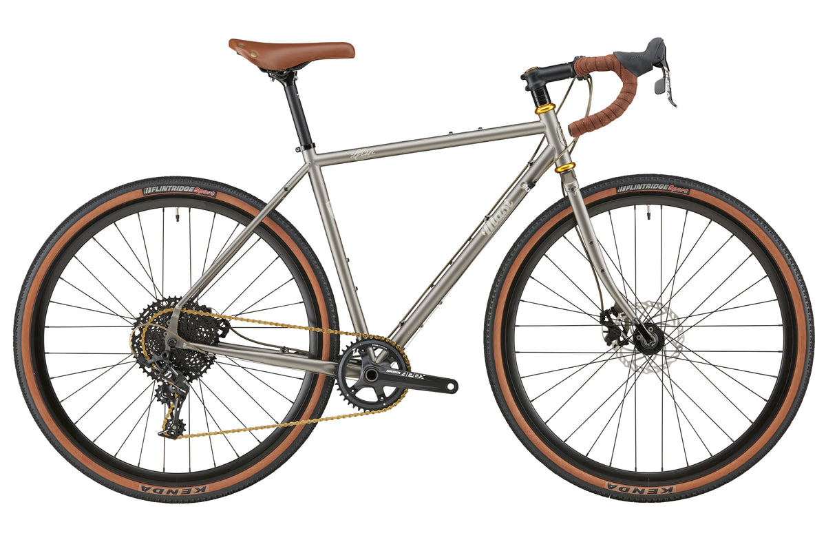 A side view of the CATALINA 1: silver gravel bike with brown handlebars, chromoly steel frame, wide tires, and disc brakes on white.
