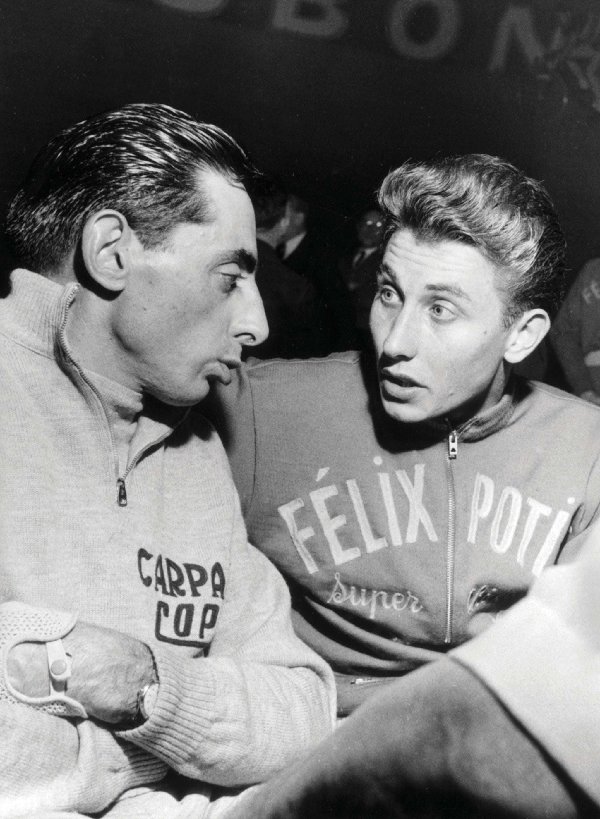 Two cyclists are engaged in conversation, both wearing sports attire with logos. One has a dark slicked-back hairstyle, and the other has light, wavy hair. The setting suggestively involves a sports-related event or discussion.