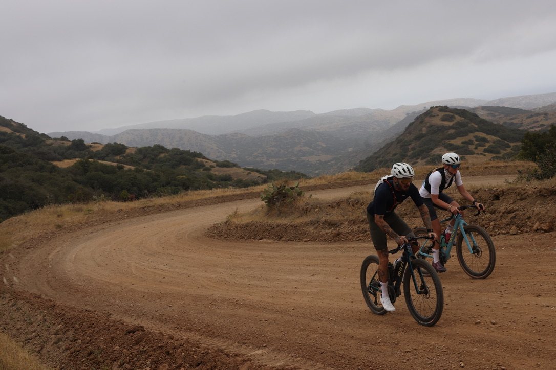 Two riders on Oltre Strada gravel bikes on a switchback dirt road above the coastal hills