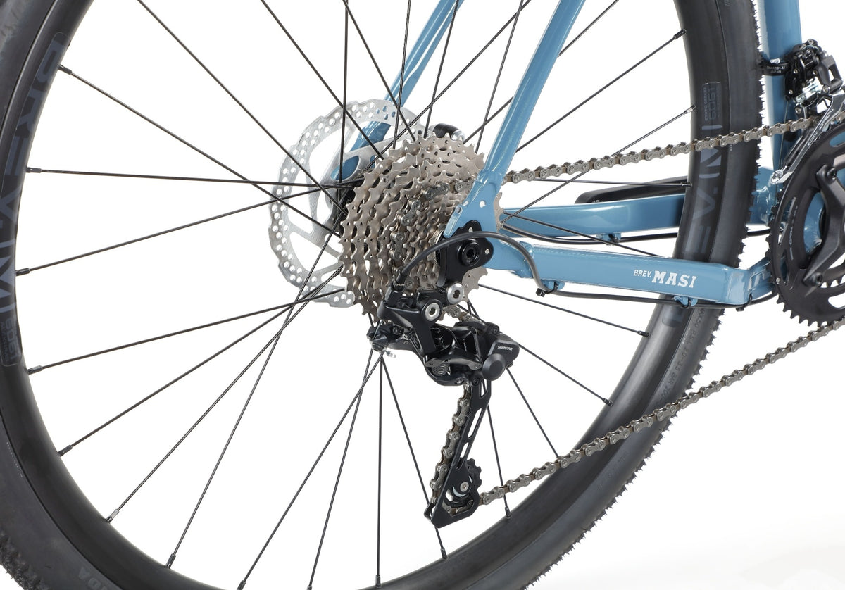 Close-up of BRUNELLO 1s rear gravel bike wheel, showing gears, derailleur, and chain on a blue alloy frame.
