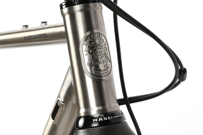 Close-up of the INCANTO CAMPY EKAR Ti bicycle frame featuring a metallic Masi Bikes badge with a crest. The brushed metal surface and black cables emphasize its sophisticated design, beautifully set against a pristine white background.